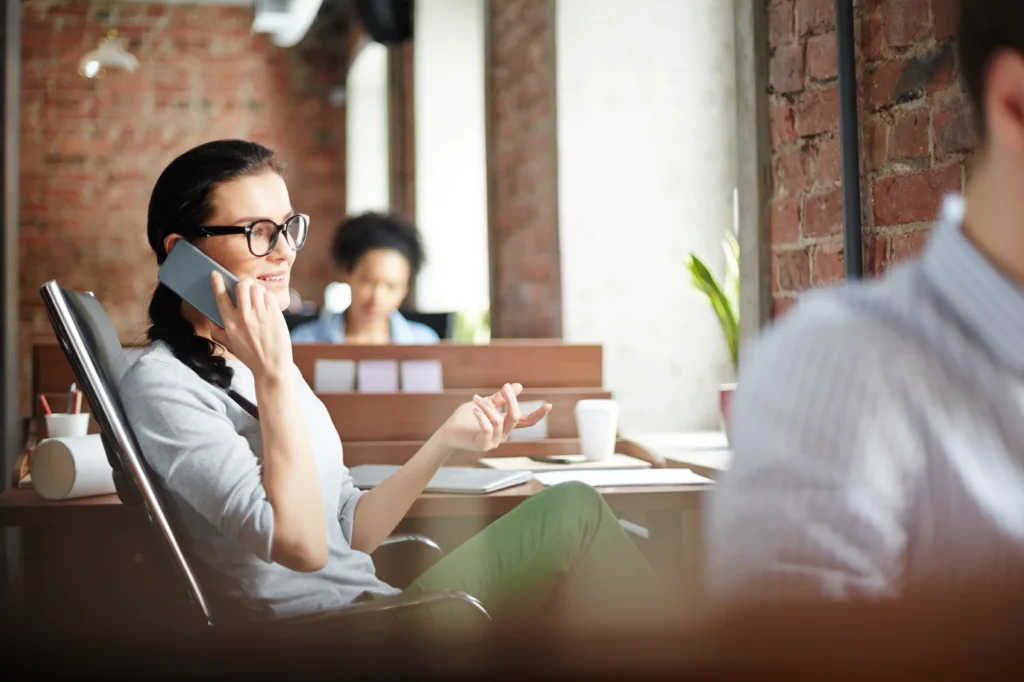 A woman in glasses talking on a mobile phone at her office desk