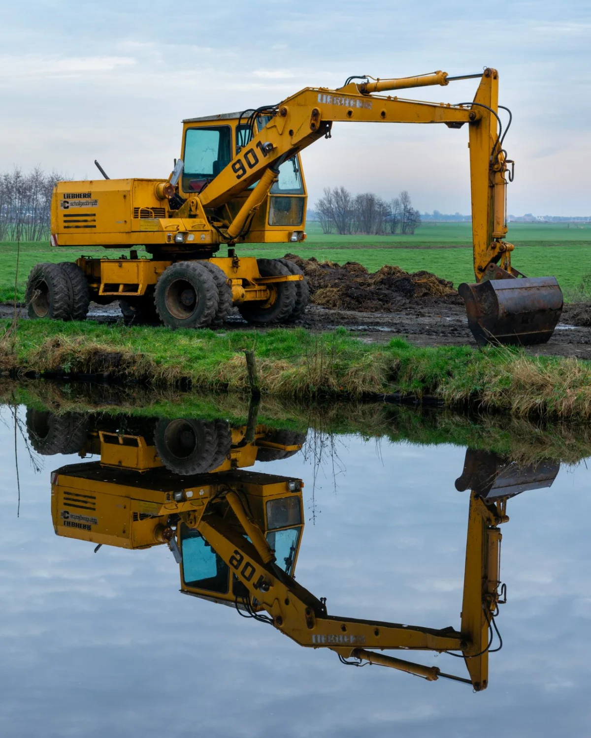 A yellow wheeled excavator digging at the edge of a calm waterway, its reflection mirrored in the water