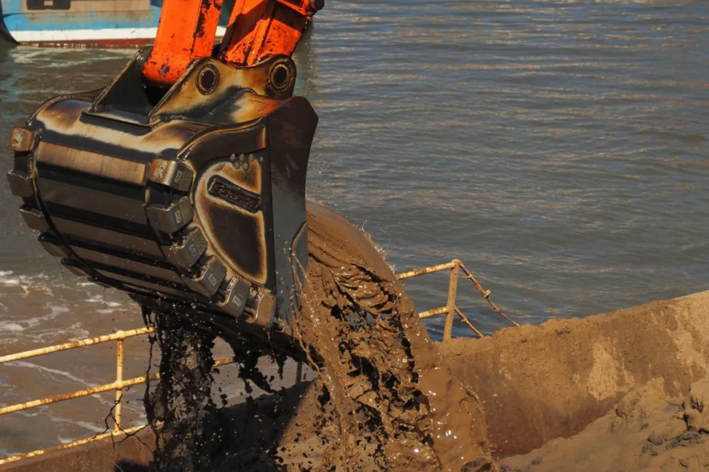 An excavator bucket lifting dredged sand and silt from the water during a harbor dredging operation