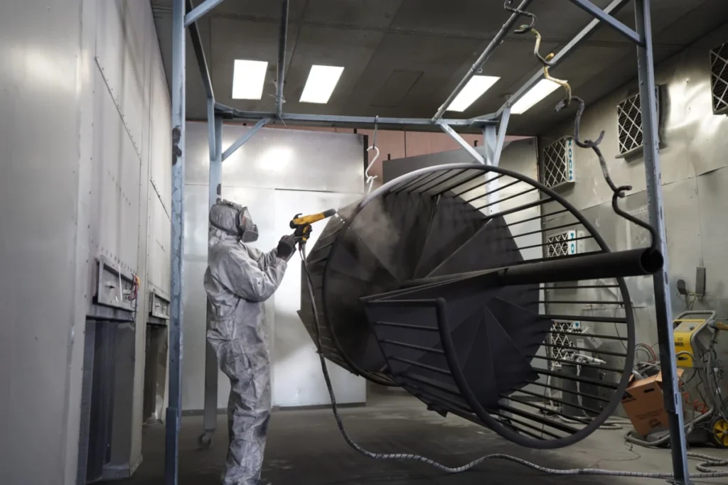 A worker in a protective suit spray-painting a large metal part in a paint booth, a next step that shapes which rust preventatives will work