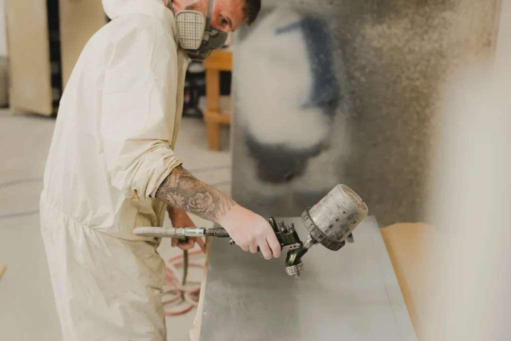A technician in protective coveralls applying a coating to a flat metal panel with a spray gun