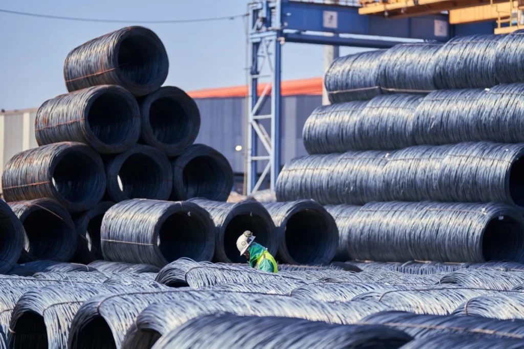 A worker walking among large bundles of coiled steel wire stacked in an industrial yard